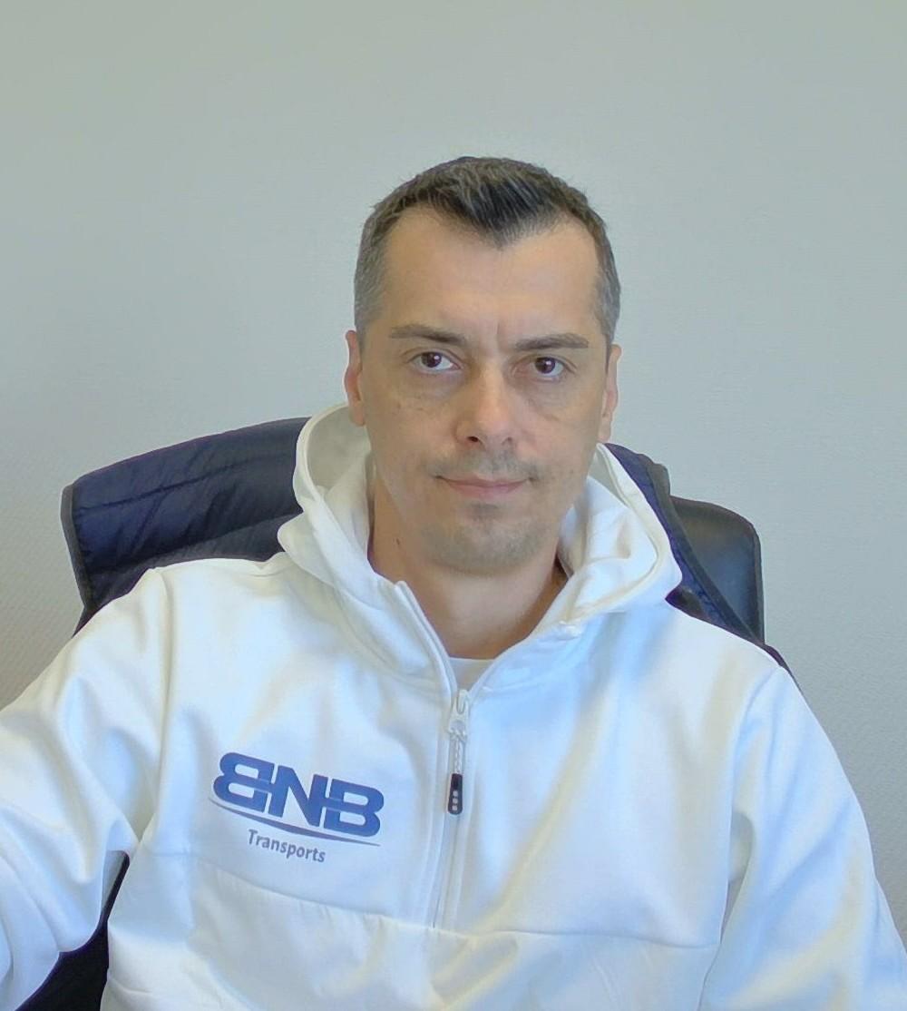 Portrait professionnel d’un dirigeant assis au bureau, portant une veste blanche avec le logo BNB Transports, sur fond clair.